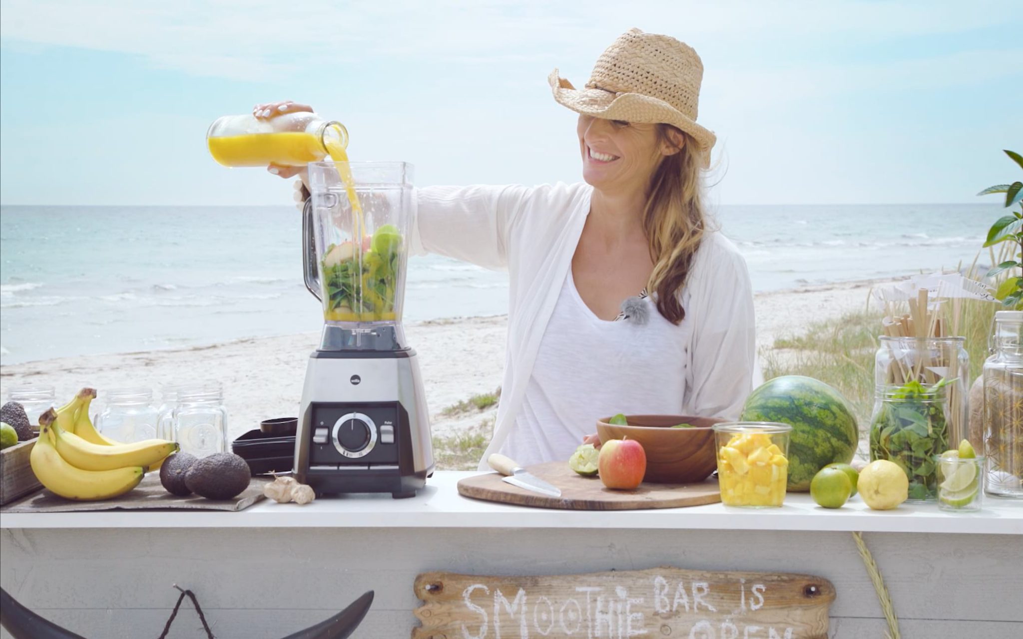 Kvinna i stråhatt står på stranden och mixar en smoothie